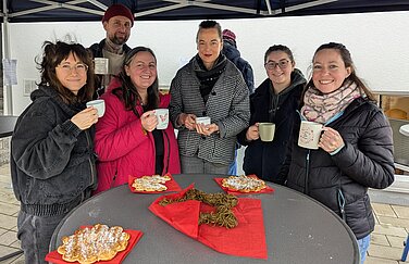 fünf Frauen und ein Mann stehen hinter einem Stehtisch, auf dem eine Waffel liegt, nebeneinander und halten Tassen mit Punsch in der Hand und blicken in die Kamera.