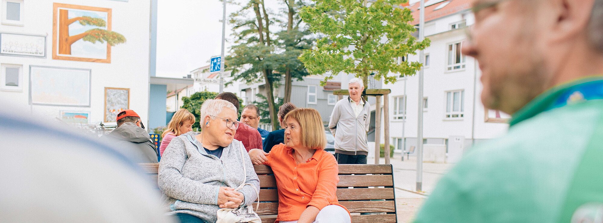 zwei Frauen sitzen auf einer Bank und unterhalten sich. im Hintergrund und auch im Vordergrund sind weitere Menschen unscharf abgebildet, die sich auf einen Vorplatz gemeinsam aufhalten.