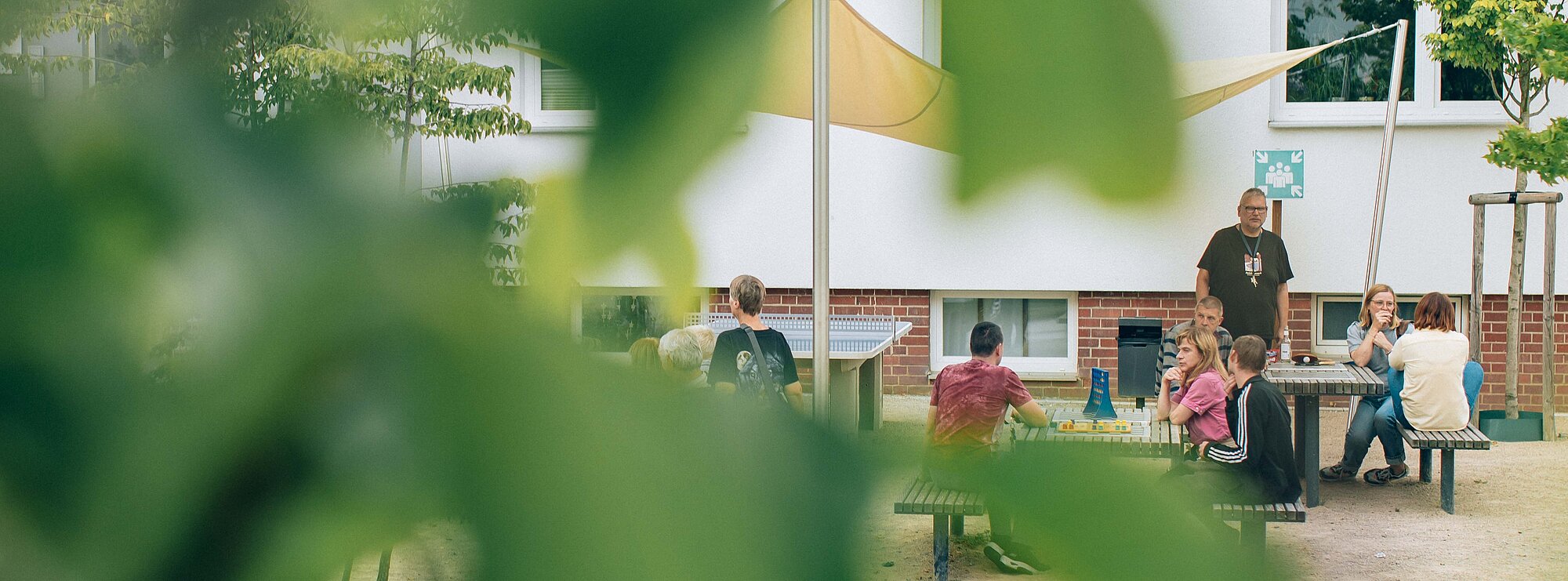 im Vordergrund sind unscharfe Blätter, durch die hindurch fotografiert wurde. Man sieht eine Gruppe von Menschen, die gemeinsam an Tischen auf Bänken sitzen und sich teilweise unterhalten.