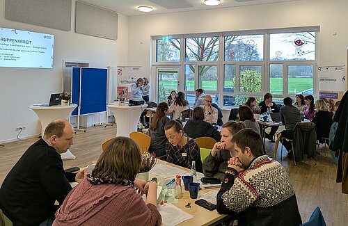 circa 30 Menschen sitzen an Gruppentischen und reden miteinander. Im Hintergrund sieht man eine große Fensterfront und mit Blick auf Felder und Bäume.