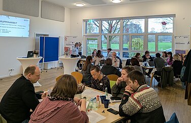 circa 30 Menschen sitzen an Gruppentischen und reden miteinander. Im Hintergrund sieht man eine große Fensterfront und mit Blick auf Felder und Bäume.