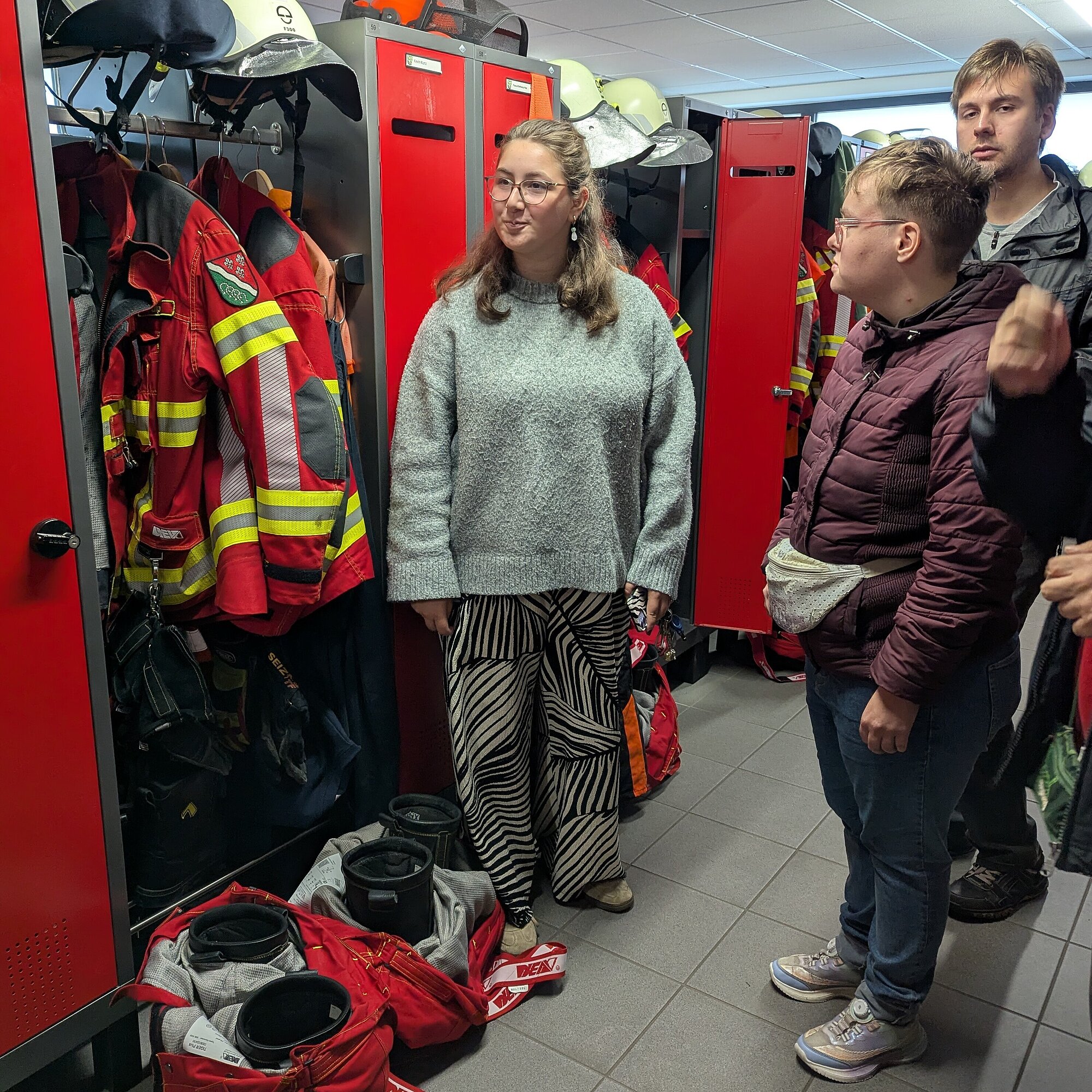 Drei Personen stehen in der Umkleide der Feuerwehr. Die Schränke sind rot. In den Schränken hängt rote Dienstkleidung.