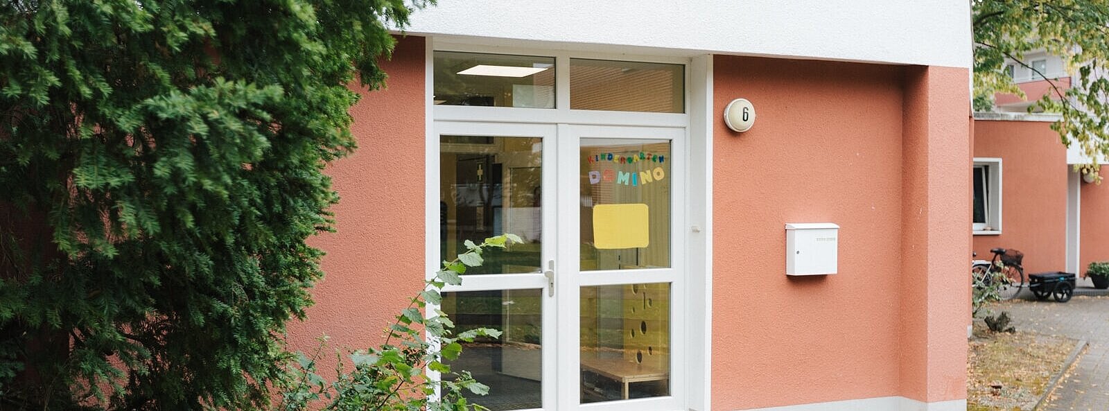 Außenansicht Kindergarten DOMINO im Böhmweg. Flachdachgebäude. Eine Glastür mit selbstgebastelter Aufschrift.