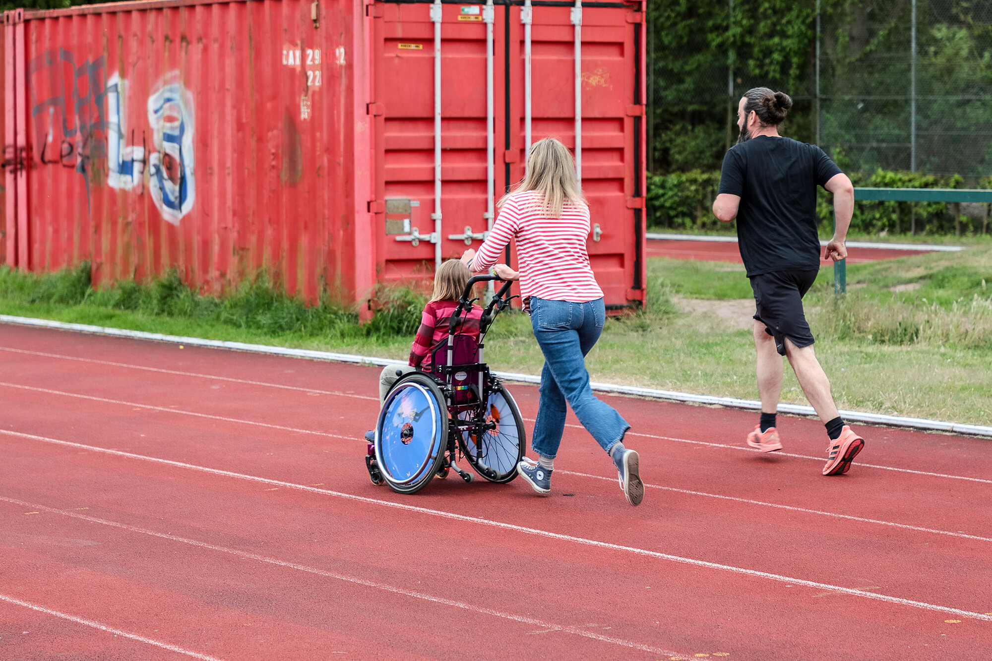 eine Frau schiebt ein Kind im Rollstuhl über die Tartanbahn, neben ihr ein Jogger