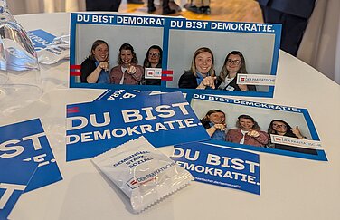 Es sind drei ausgedruckte Bilder zu sehen. Auf diesen Bildern befinden sich drei Personen, die mit einem ernsten Blick zur Kamera gucken und dabei den Finger zur Kamera richten. Die Ausdrucke, Postkarten und kleine Pakete mit Gummibärchen tragen die Aufschrift "Du bist Demokratie"