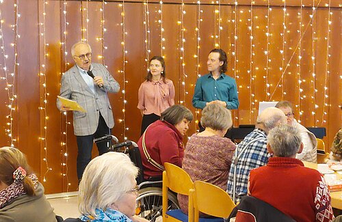 Der Vorsitzende Rainer Weidler steht neben einer Frau und einem Mann und hält eine Rede. Vor den dreien sitzen Gäste an Tischen. Sie schauen zum Redner und man sieht sie nur von hinten.