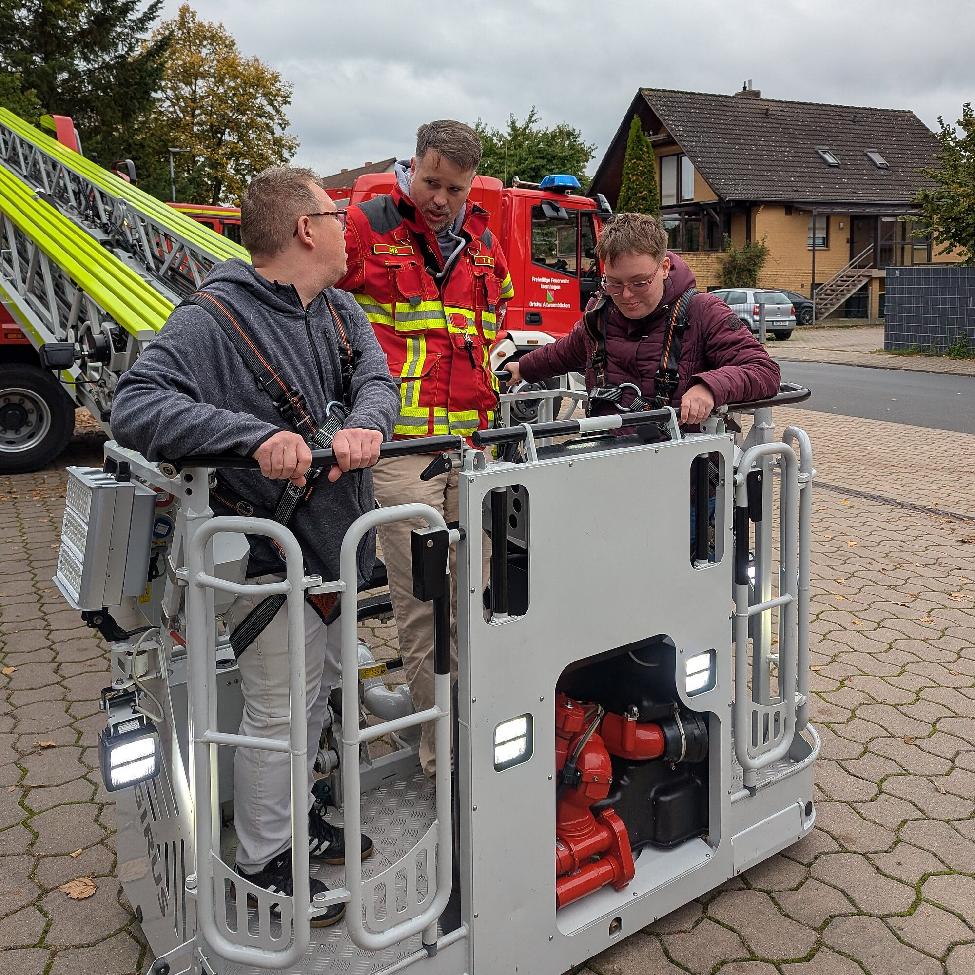 Drei Personen stehen im Korb einer Drehleiter. Eine Person davon ist Feuerwehrmann. Sie werden mit der Drehleiter hoch fahren