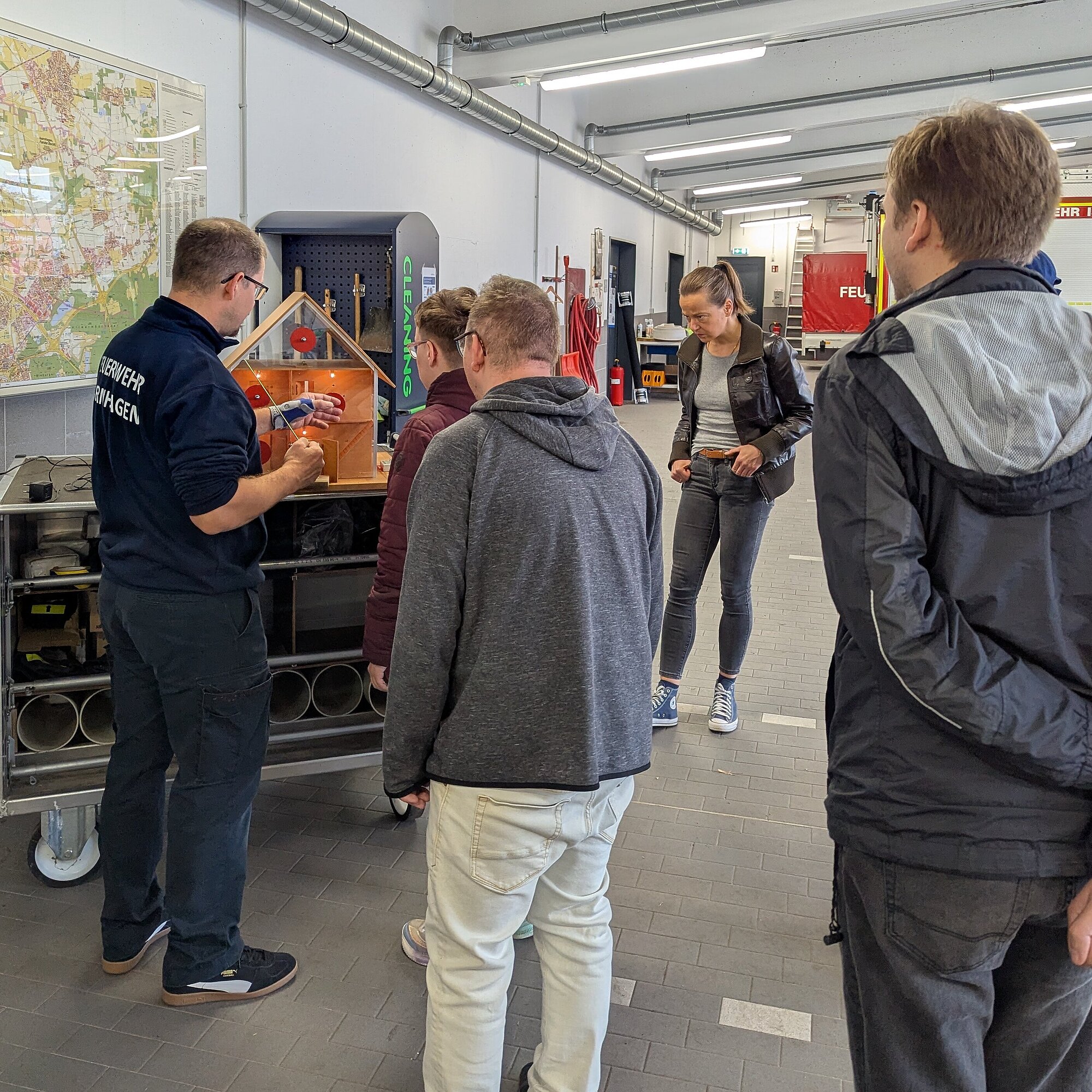 Vier Personen stehen in einer Fahrzeughalle der Feuerwehr. In einem kleinen Glashaus wird die Ausbreitung von Rauch simuliert