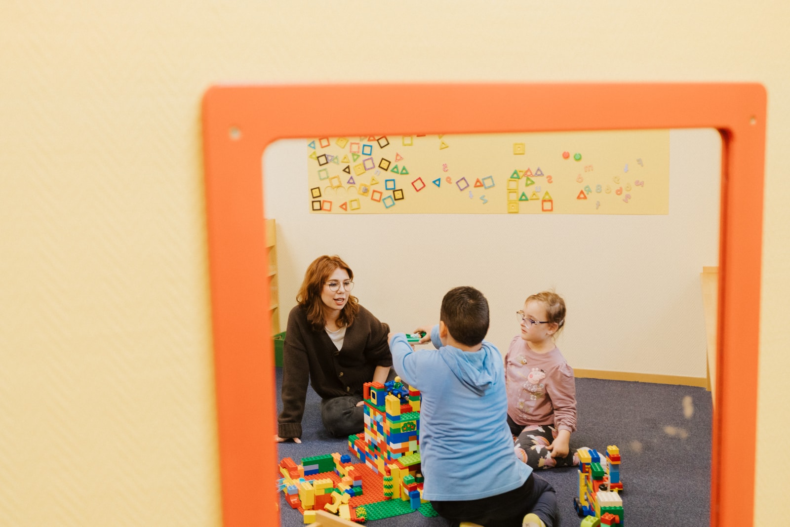 Es spiegeln sich drei Personen in einem Spiegel. Sie sitzen gemeinsam auf einem Teppich und spielen zusammen mit Bauklötzen