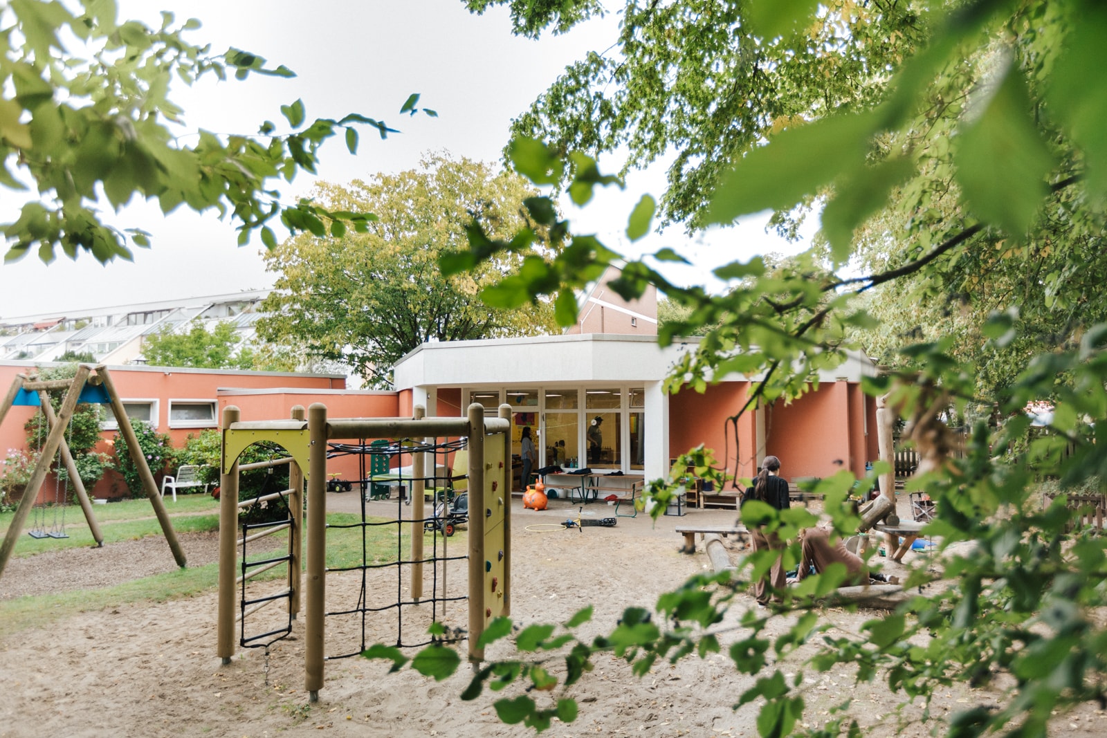 Außenbereich des Kindergarten DOMINO. Im Vordergrund sind Büsche, ein Klettergerüst und im Hintergrund der Kindergarten