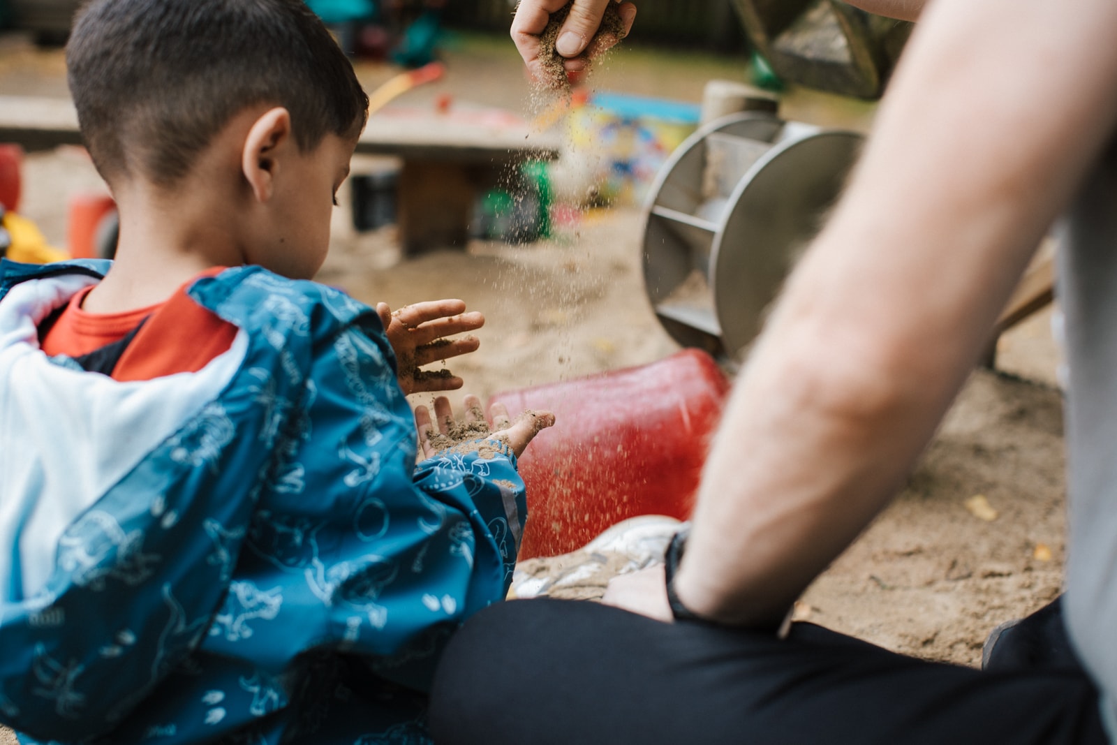 Kind von hinten im Sandkasten sitzend. Man sieht einen Arm eines Erwachsenen der Sand aus der Hand rieseln lässt. 
